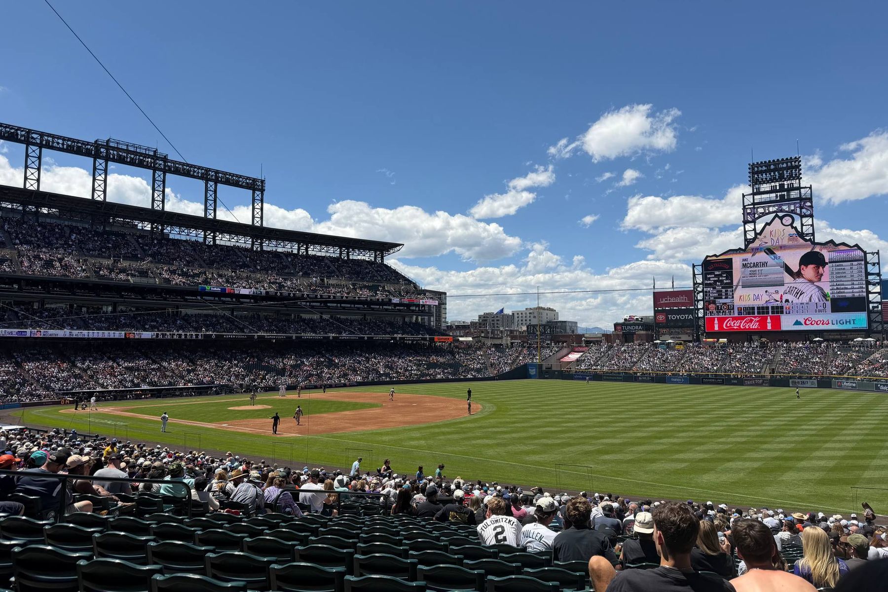 Coors Field during a day game, the full lower bowl and outfield seen from an elevated seat behind home plate under a blue Colorado sky