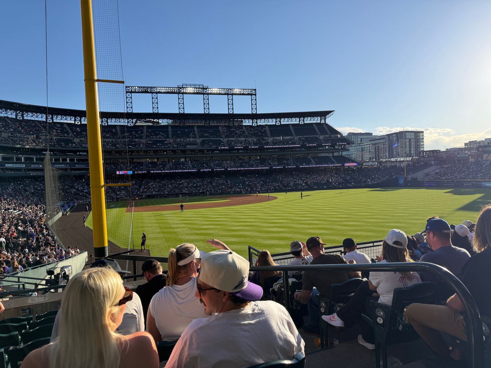 The view from the right-field corner seats at Coors Field behind the yellow foul pole at a night game, the setting sun low over the grandstand and flaring into the frame
