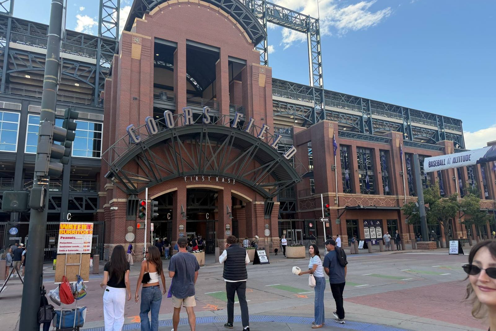 Fans walking across the plaza toward the arched main entrance at Coors Field, the COORS FIELD sign above the gate