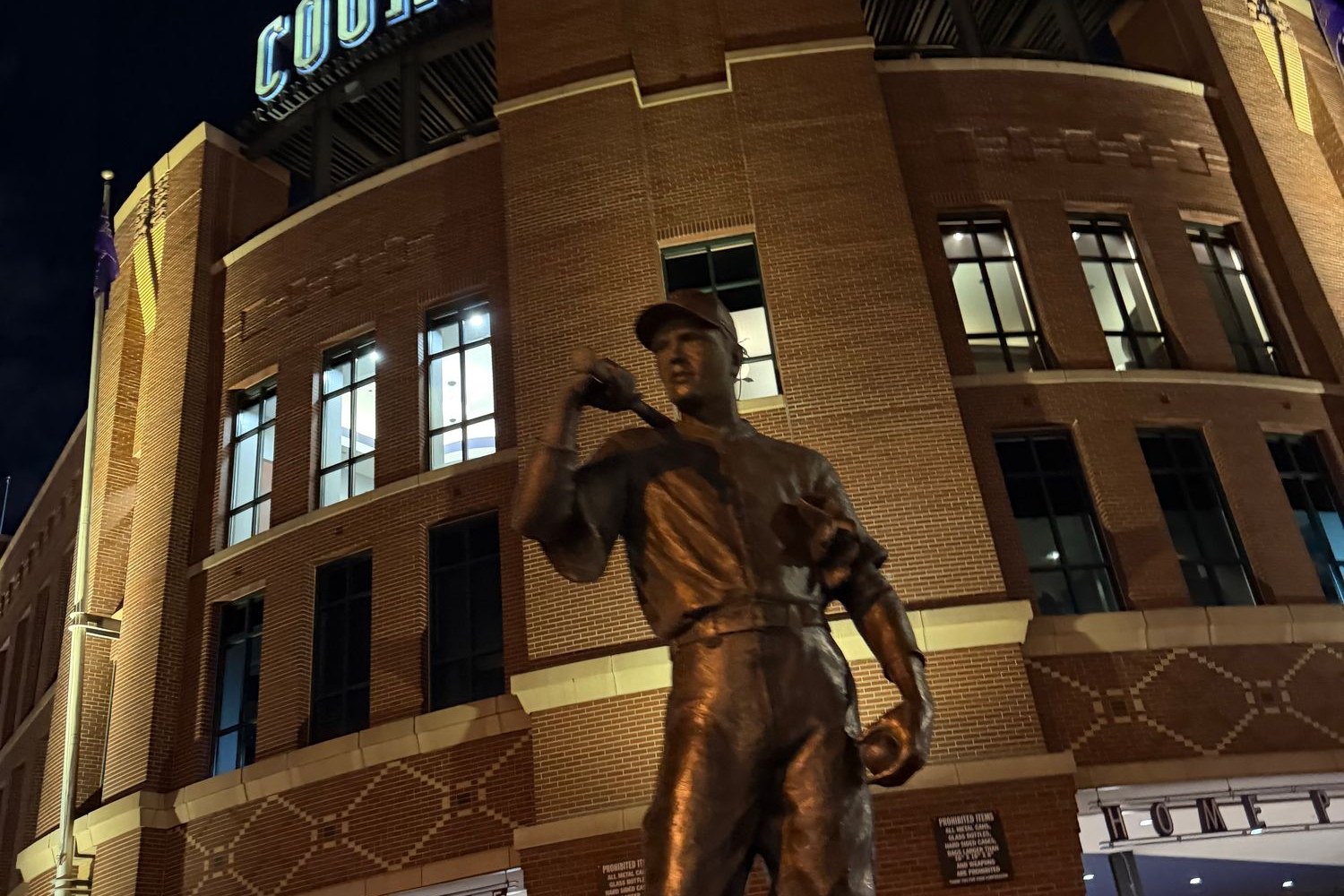 A bronze statue of a baseball player outside Coors Field, lit against the ballpark's brick facade at night