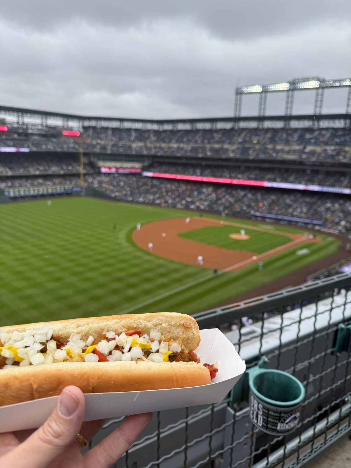 A loaded hot dog topped with onions and mustard held up in a paper tray at Coors Field, the field and lower bowl visible beyond an upper-level rail