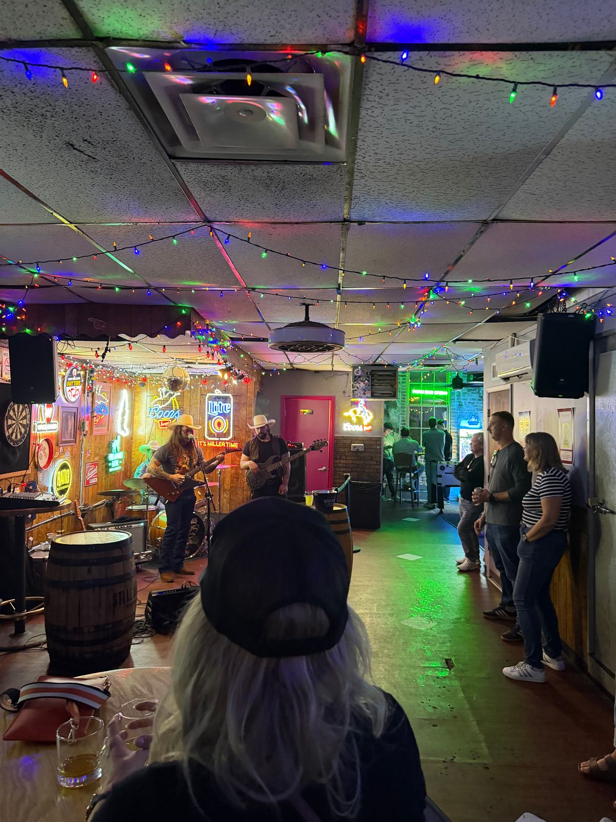The interior of Star Bar in Denver on a live-music night, a band playing under string lights and neon beer signs, whiskey barrels and a dartboard along the walls