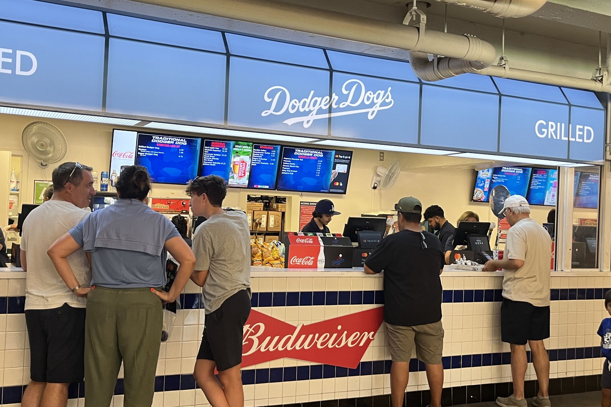 The Dodger Dogs stand on the concourse at Dodger Stadium, GRILLED lettered on the blue awning beside the script sign, menu boards lit above the counter and fans lined up along the tiled rail