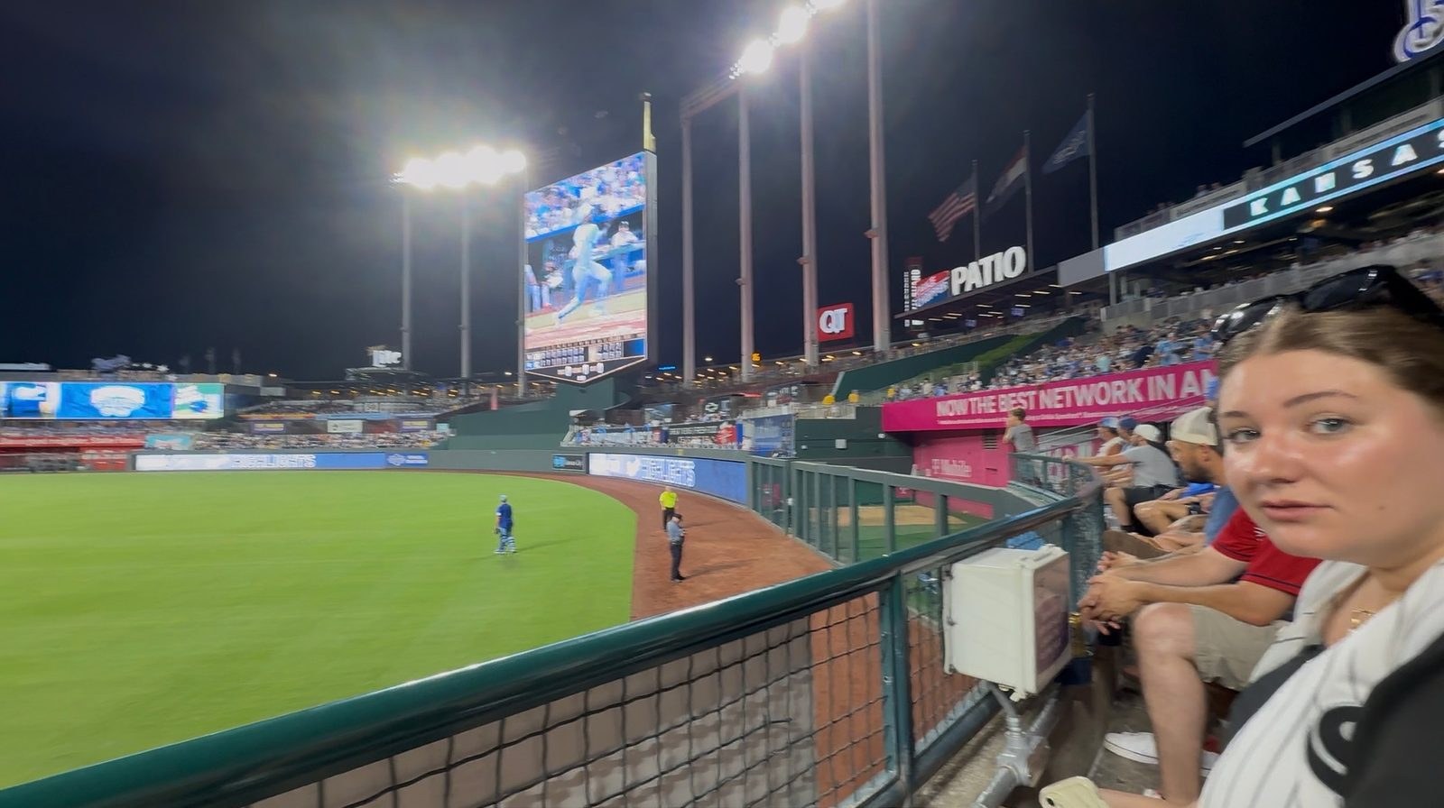 A night view from the outfield patio at Kauffman Stadium looking in toward the lit crown scoreboard, with the bowl glowing under the lights and fans along the rail