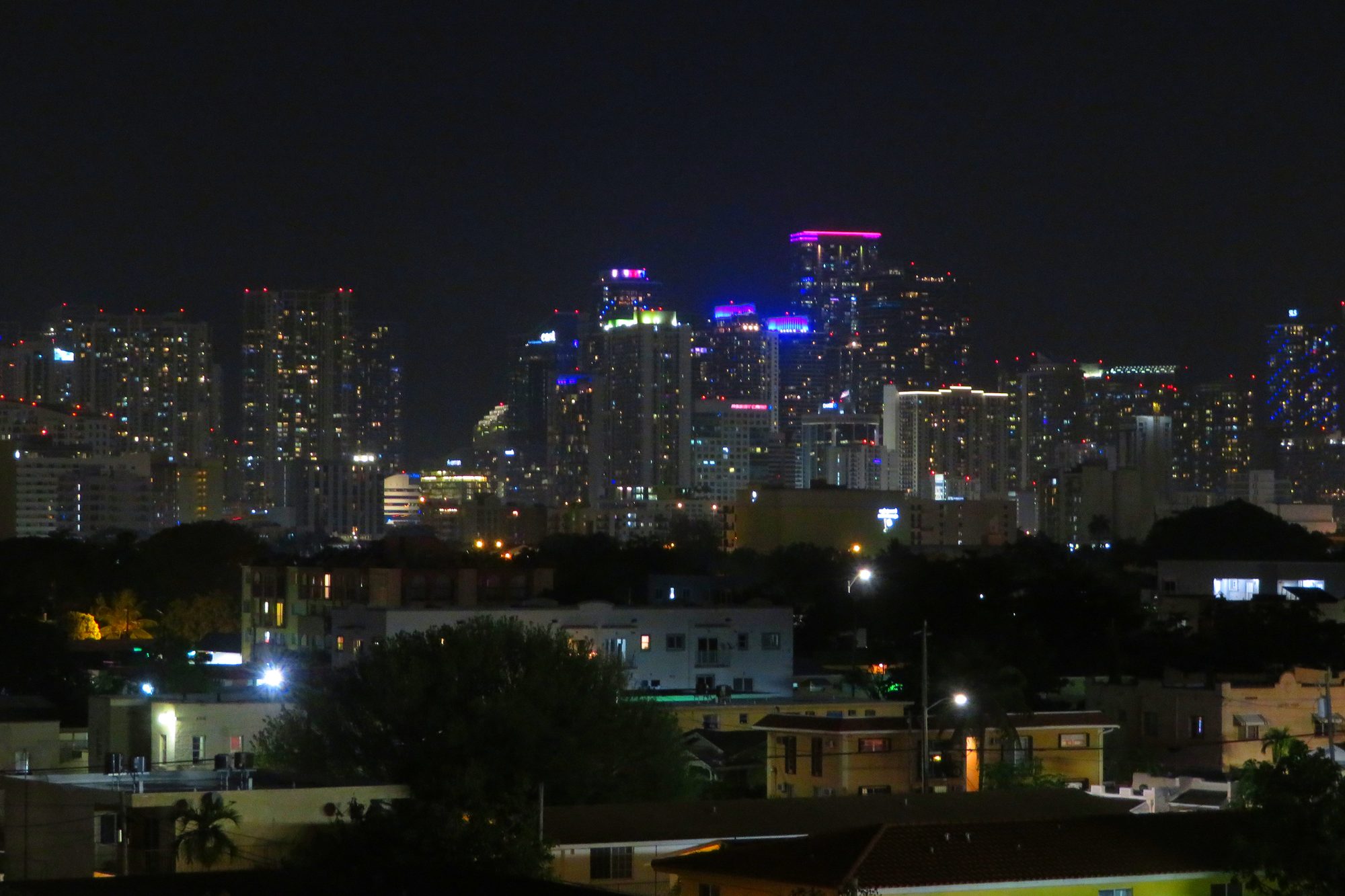 The downtown Miami skyline lit up at night, its towers rising beyond the low rooftops and palms of Little Havana, seen from loanDepot park