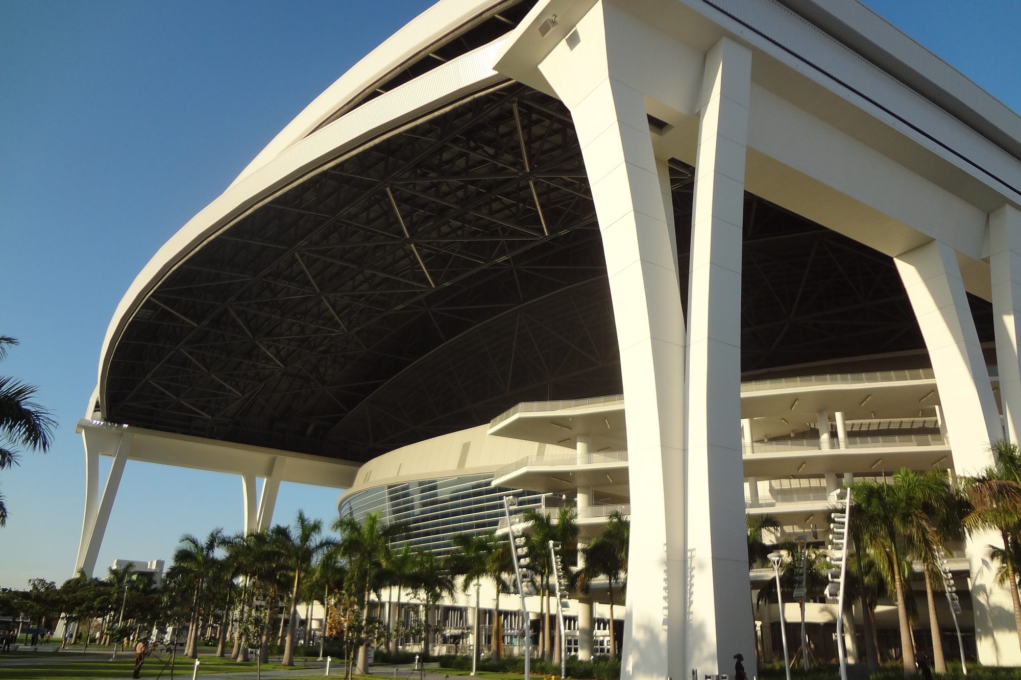 The underside of the retractable roof at loanDepot park, its steel lattice and the white structural columns that carry it, seen from the plaza against a clear Miami sky