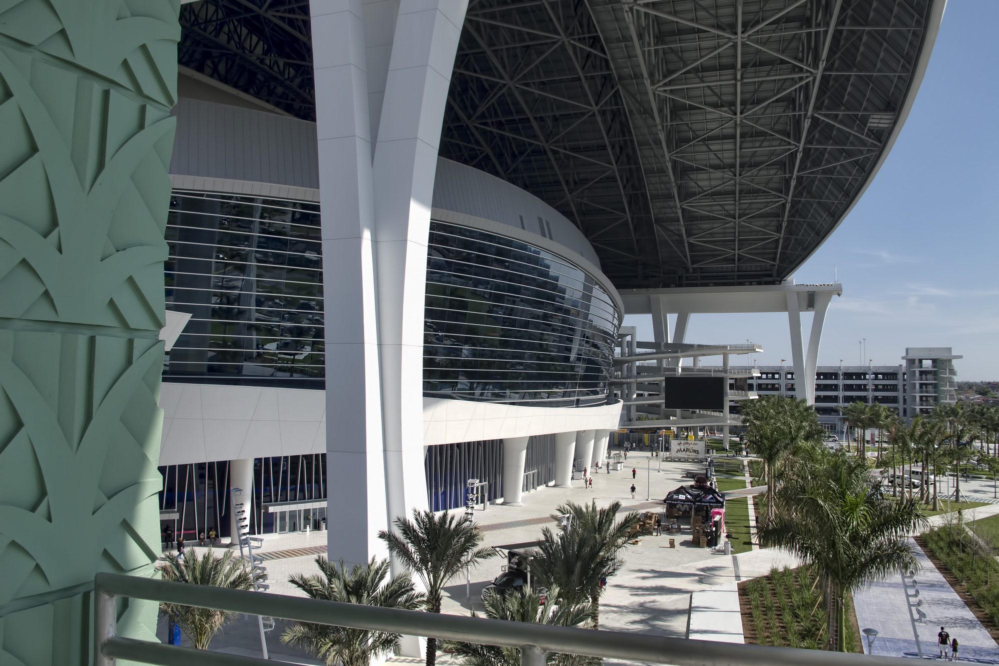 The West Plaza at loanDepot park, fans crossing the open plaza beneath the cantilevered roof, the curved glass concourse wall and the pale green perforated facade panel framing the approach