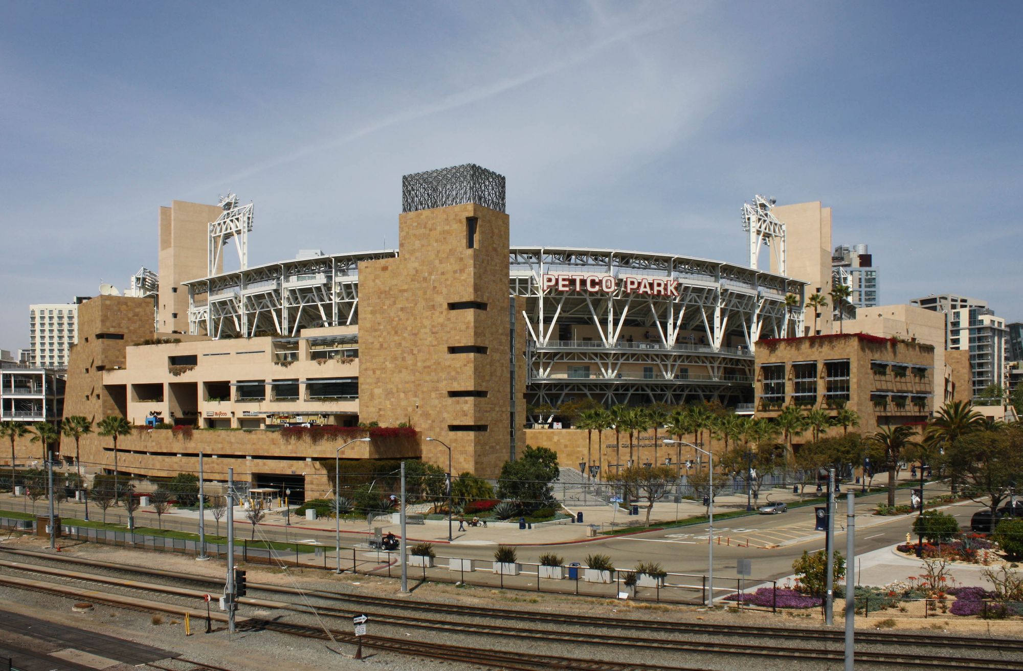 The sandstone exterior of Petco Park from across the trolley tracks on a clear day, PETCO PARK lettering on the facade beneath the white light towers, with palm trees lining the street below
