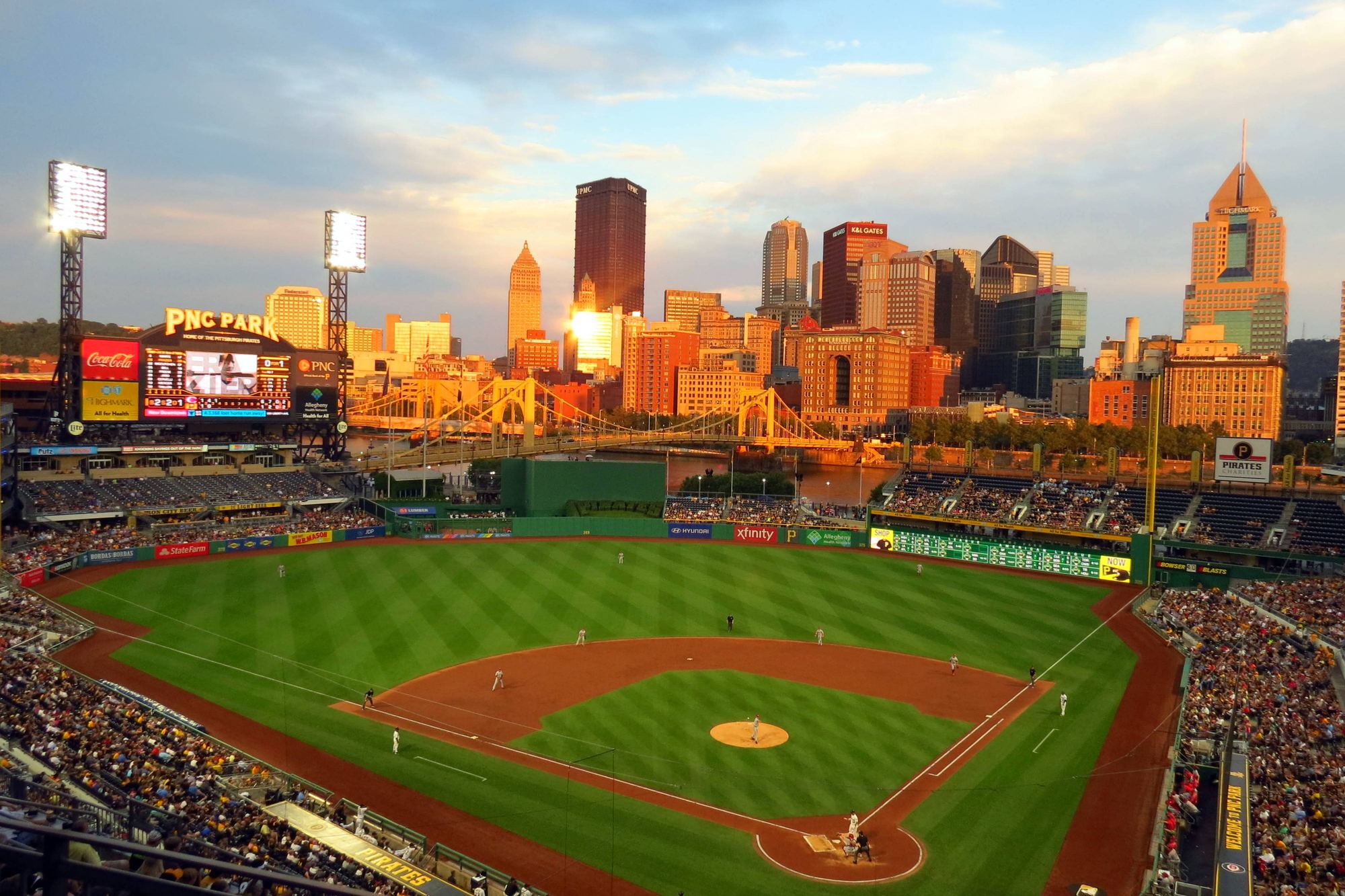 PNC Park at golden hour, the sun setting behind the downtown Pittsburgh skyline beyond the outfield