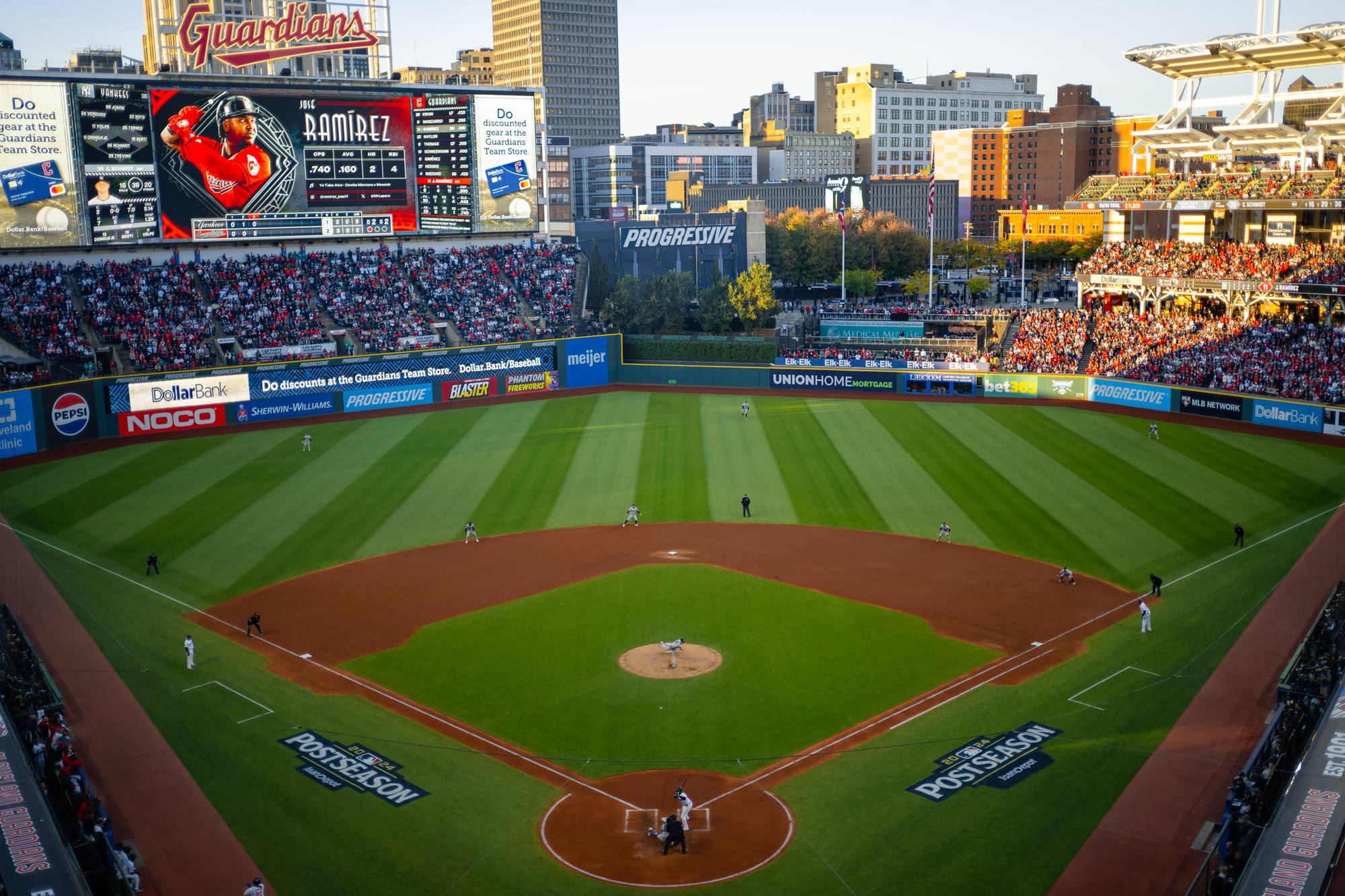 Progressive Field packed for a 2024 postseason game against the Yankees, seen from high behind home plate, MLB postseason logos on the grass and the downtown Cleveland buildings beyond the outfield