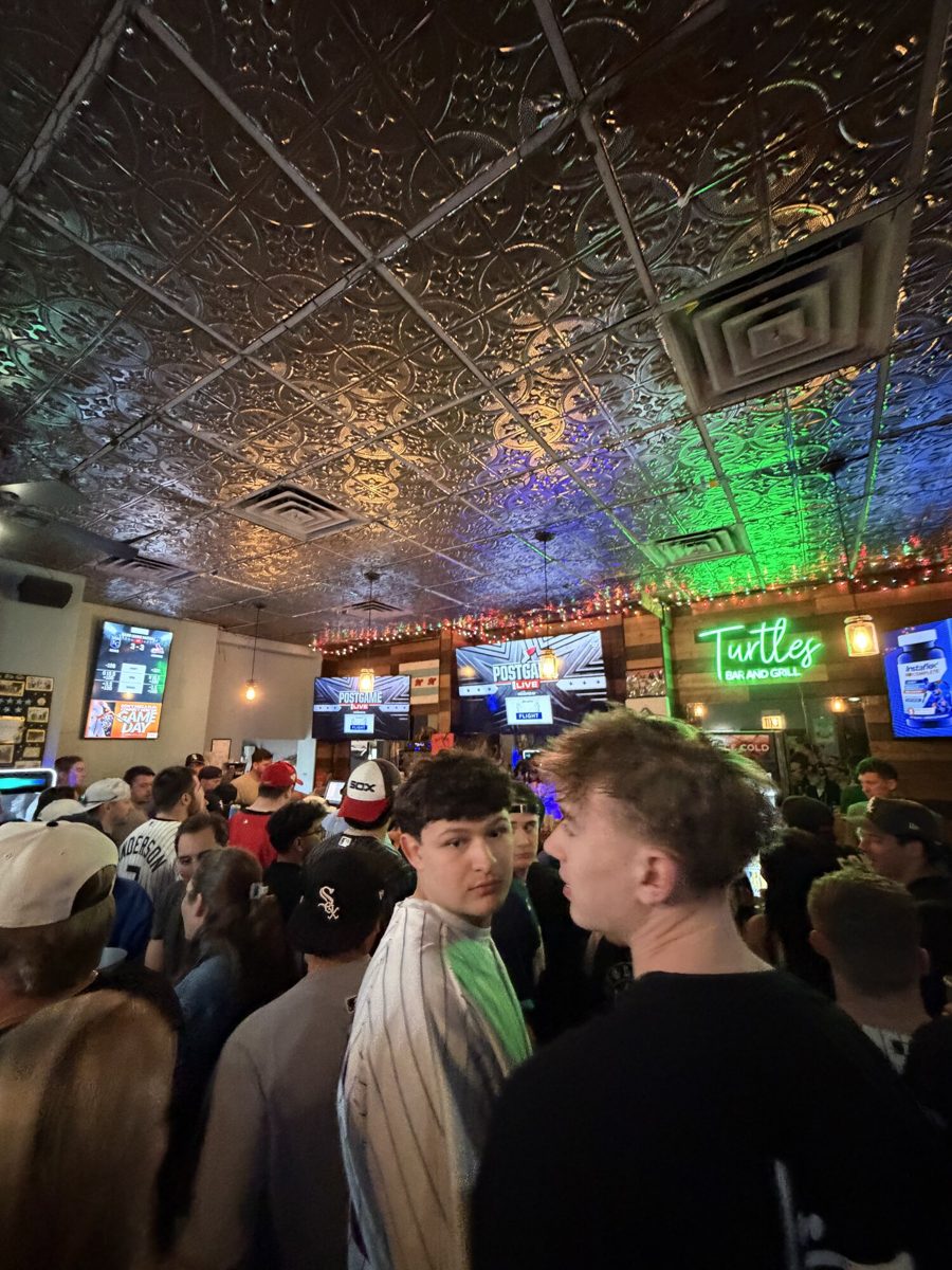 The interior of Turtle's Bar & Grill at night, with the green Turtle's neon sign glowing above an old pressed-tin ceiling and a crowd of Sox fans packed at the bar