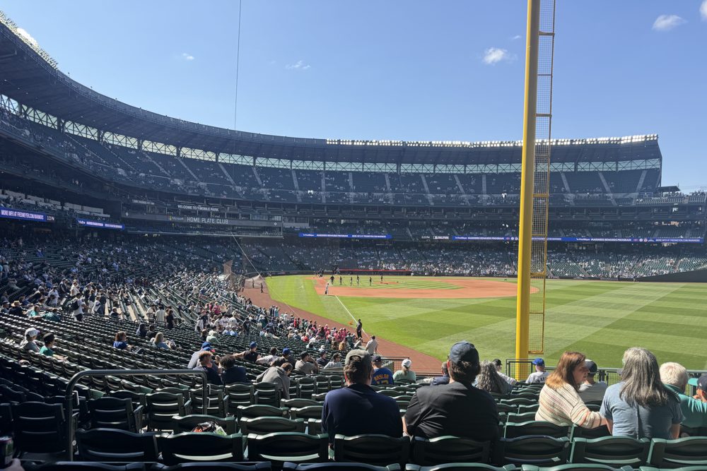 Fans in the 100-level seats at T-Mobile Park looking down the line toward a yellow foul pole, with the stands across the field on a sunny day