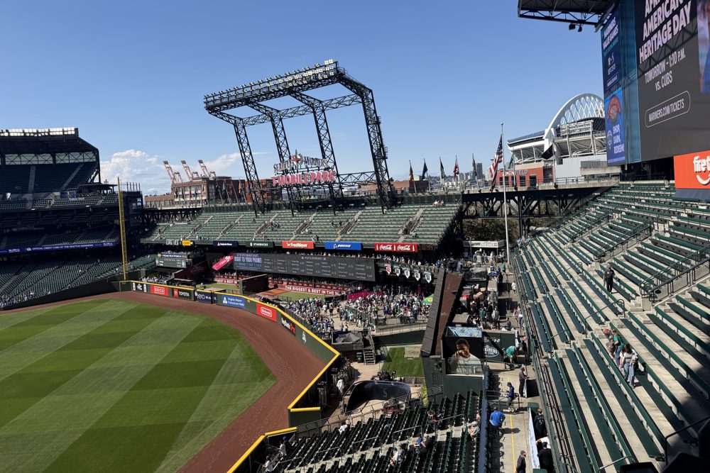 The left field bleachers and outfield at T-Mobile Park under a clear sky, with the roof's rail structure and the port cranes beyond