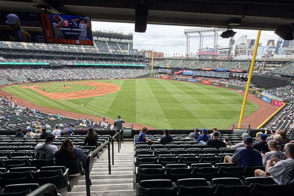 The field at T-Mobile Park seen from the back of a covered section, with the overhang and a mounted TV across the top of the frame and sunlit rows below