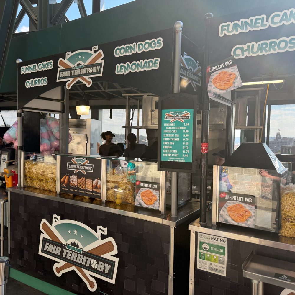 The Fair Territory stand at T-Mobile Park with signs for churros, funnel cakes, corn dogs, and lemonade, and cotton candy and popcorn in the window