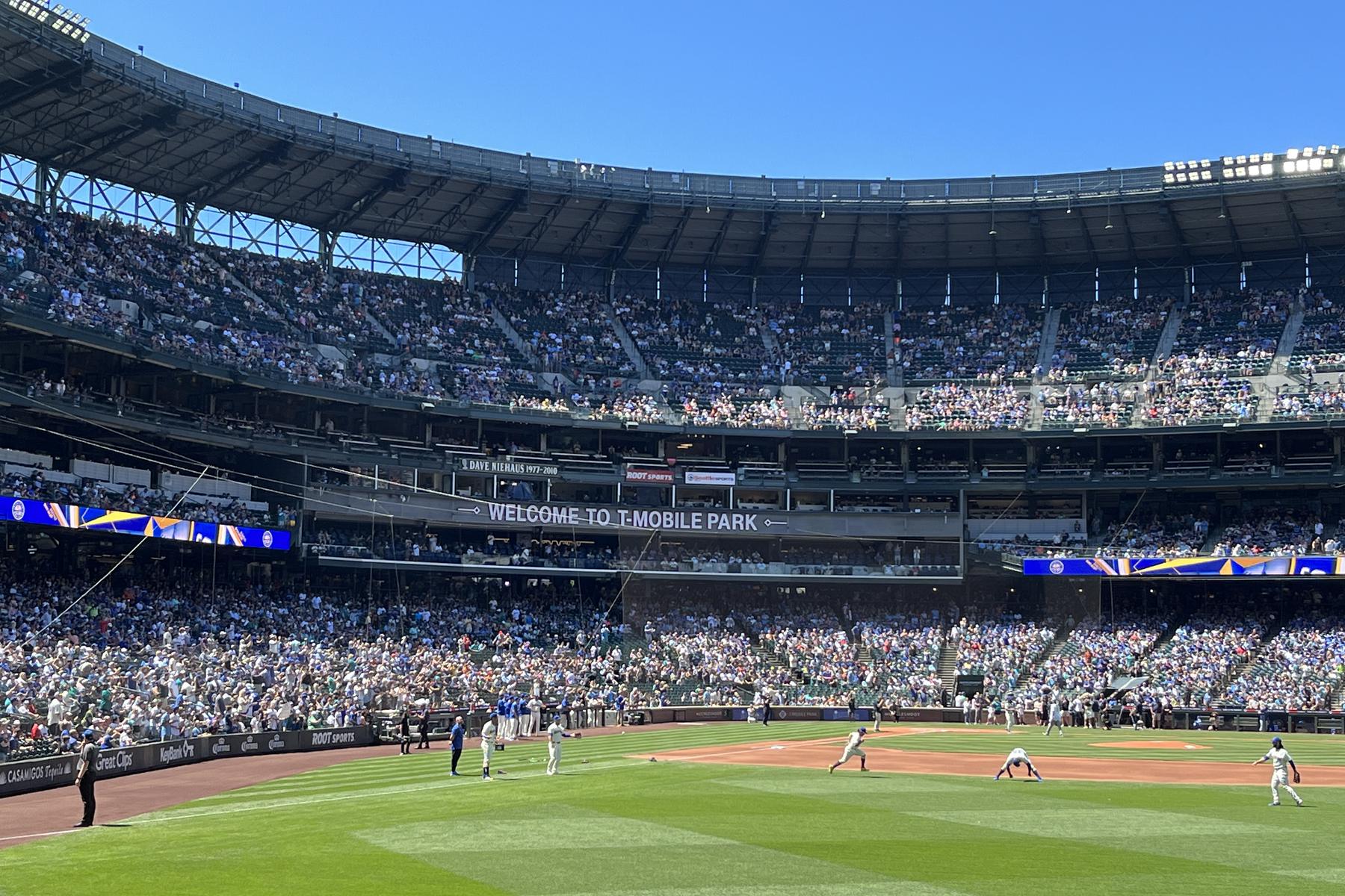 A packed T-Mobile Park on a clear summer day, a banner across the left-field deck facade reading Welcome to T-Mobile Park, the roof structure overhead and the game underway