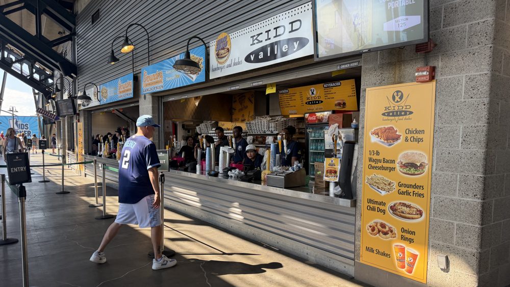 The Kidd Valley stand on a sunny concourse at T-Mobile Park, its yellow menu poster listing burgers, chicken and chips, and shakes, with one fan walking past