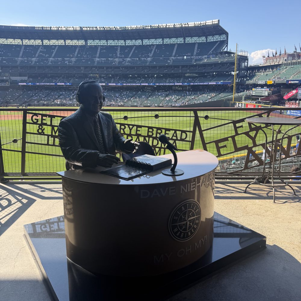 The bronze Dave Niehaus statue seated at a broadcast desk inside T-Mobile Park, backlit, with the field and the outfield seats behind