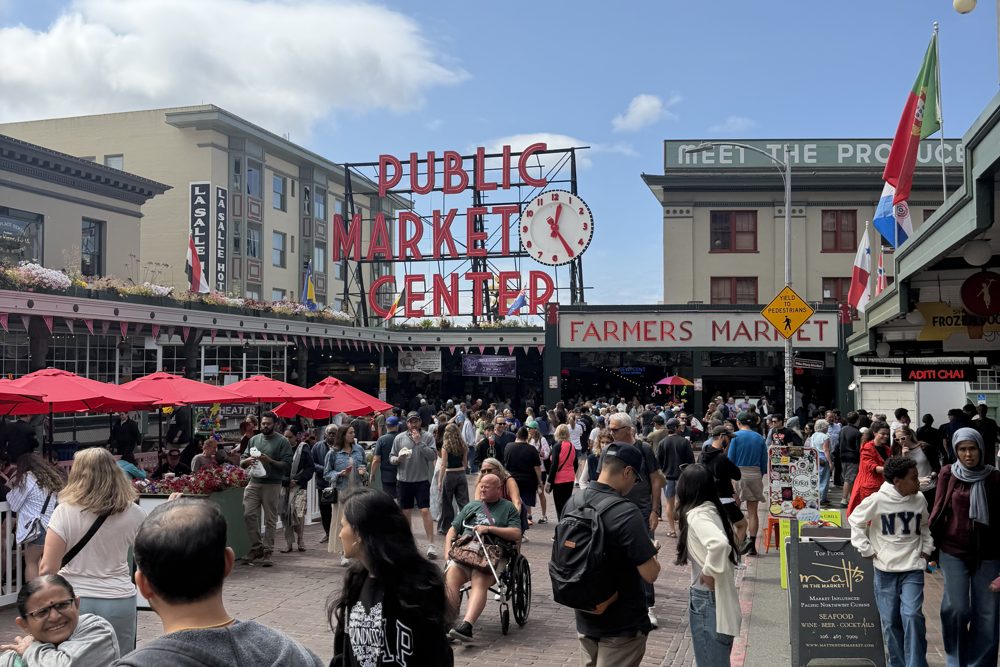 The Public Market Center sign and clock at Pike Place Market in Seattle, with red umbrellas and a busy crowd below on a sunny day