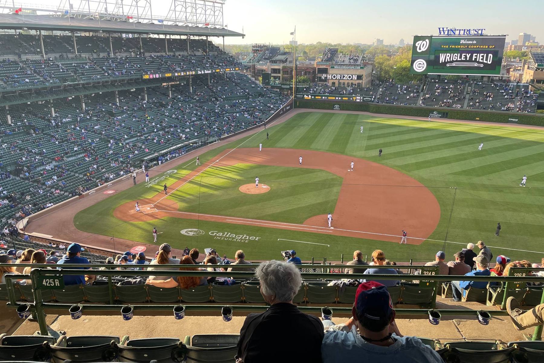 The field at Wrigley Field seen from an upper-deck seat in golden evening light, fans watching from the rows below