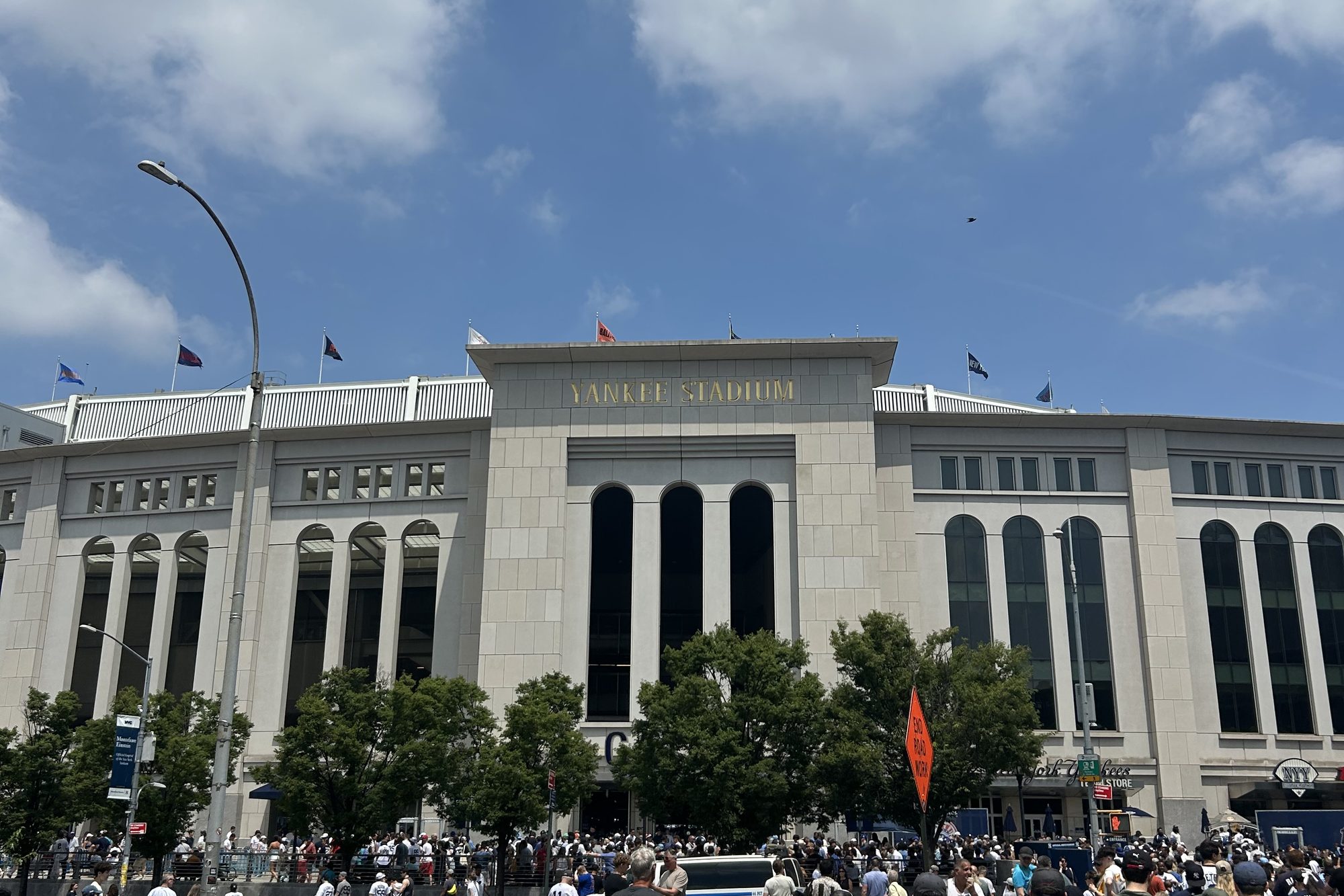 The limestone facade of Yankee Stadium on a game day, YANKEE STADIUM in gold lettering above the arched windows, flags along the roofline and a crowd filling the sidewalk outside the team store